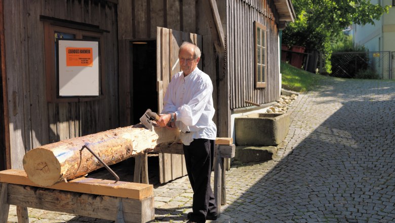 Lebendiges Handwerk, © FVV Ein Mann bearbeitet einen Baumstamm mit einer Axt vor einem Holzhaus.