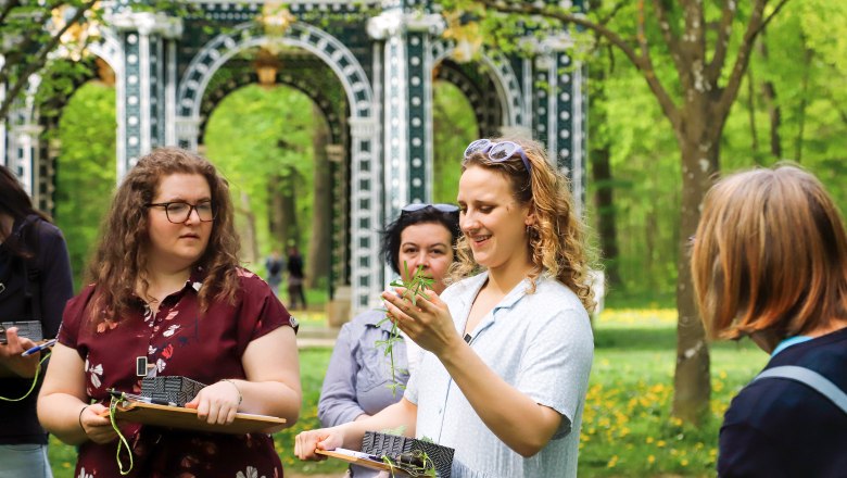 Eine Gruppe von Menschen bei einem Kr&auml;uterworkshop im Freien, umgeben von B&auml;umen und einem dekorativen Torbogen.
