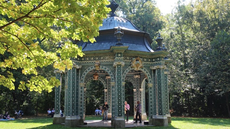 Ein gr&uuml;nes Lusthaus im Park, umgeben von B&auml;umen und Menschen.