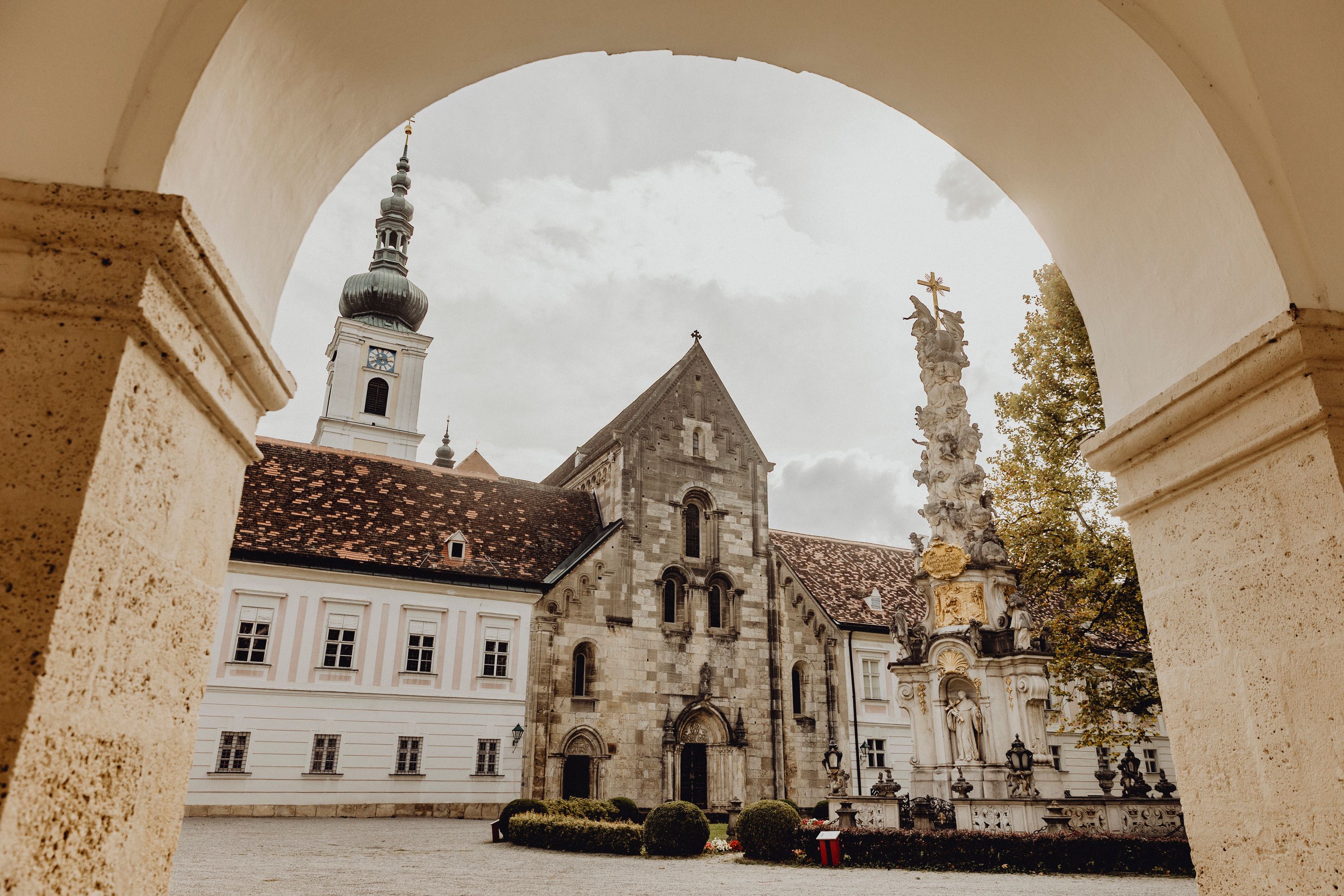 Blick durch einen Torbogen auf ein historisches Klostergebäude mit Turm und einer barocken Säule im Vordergrund.