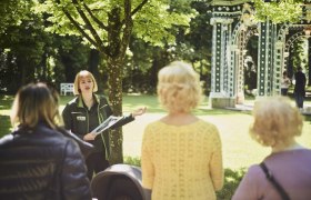 Eine Frau f&uuml;hrt eine Gruppe durch einen sonnigen Park mit einem dekorativen Pavillon im Hintergrund.