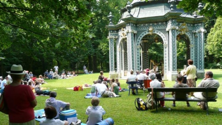 Menschen sitzen auf einer Wiese vor einem dekorativen Pavillon im Gr&uuml;nen.