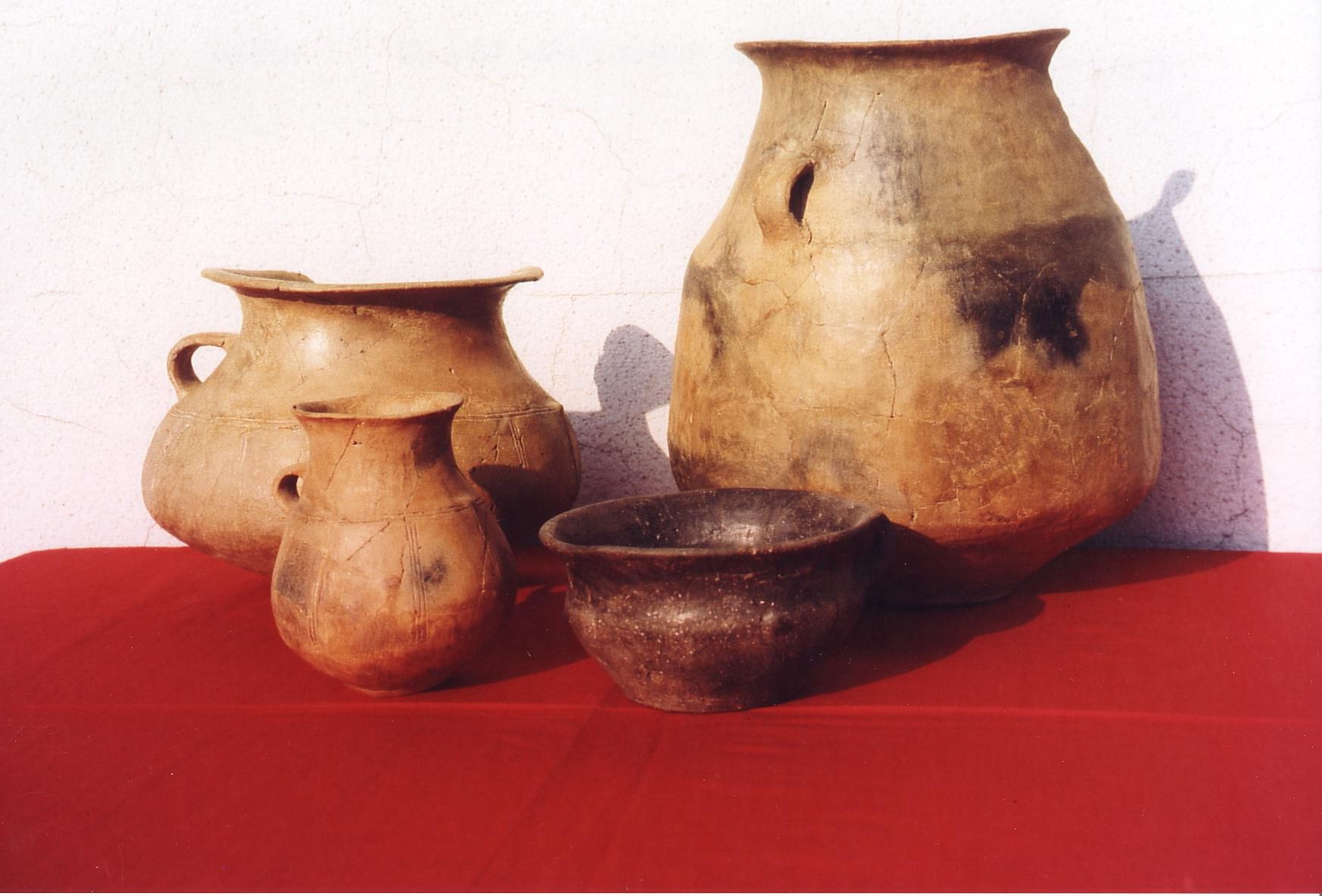 Four antique clay vessels on red cloth in front of a white wall.