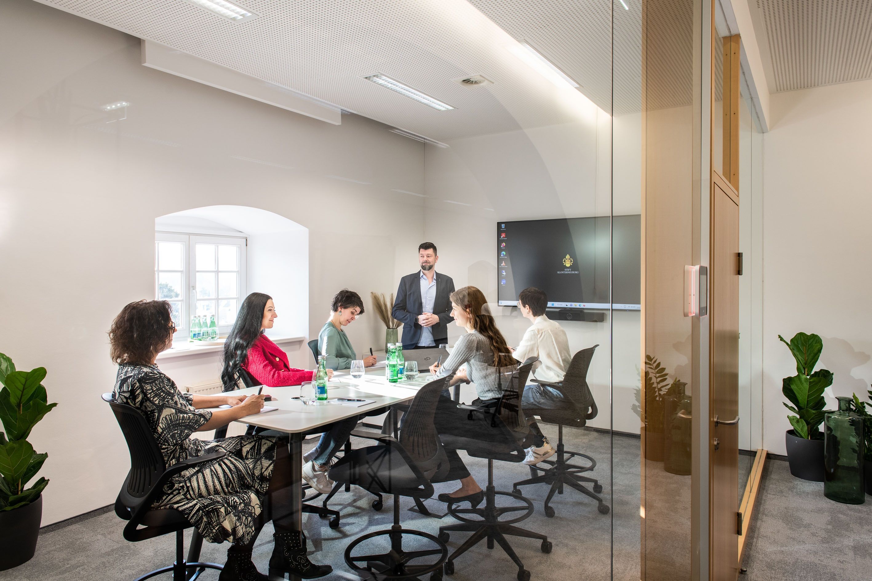 A modern conference room with six people sitting at a table listening to a speaker. A screen in the background.