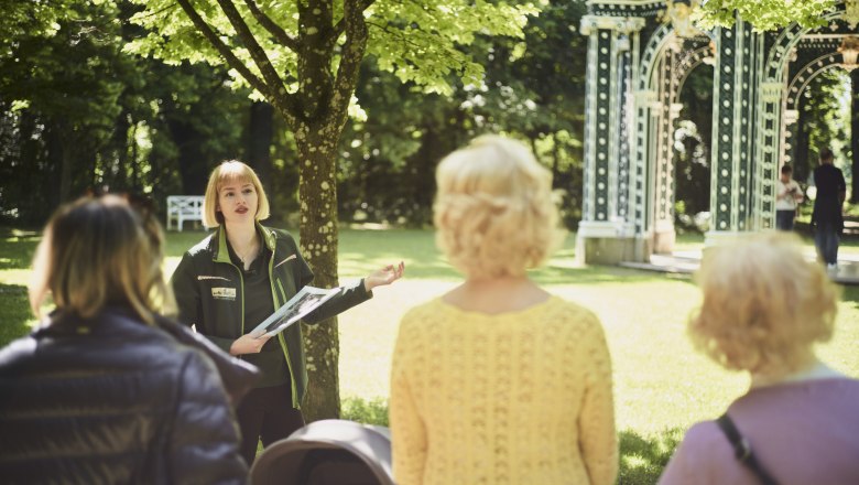 Eine Frau f&uuml;hrt eine Gruppe durch einen sonnigen Park mit einem dekorativen Pavillon im Hintergrund.