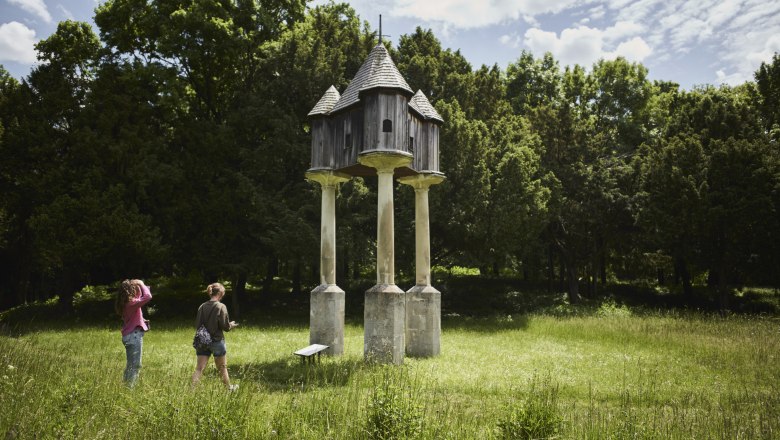 Zwei Personen betrachten eine h&ouml;lzerne Struktur auf S&auml;ulen in einem gr&uuml;nen Park.