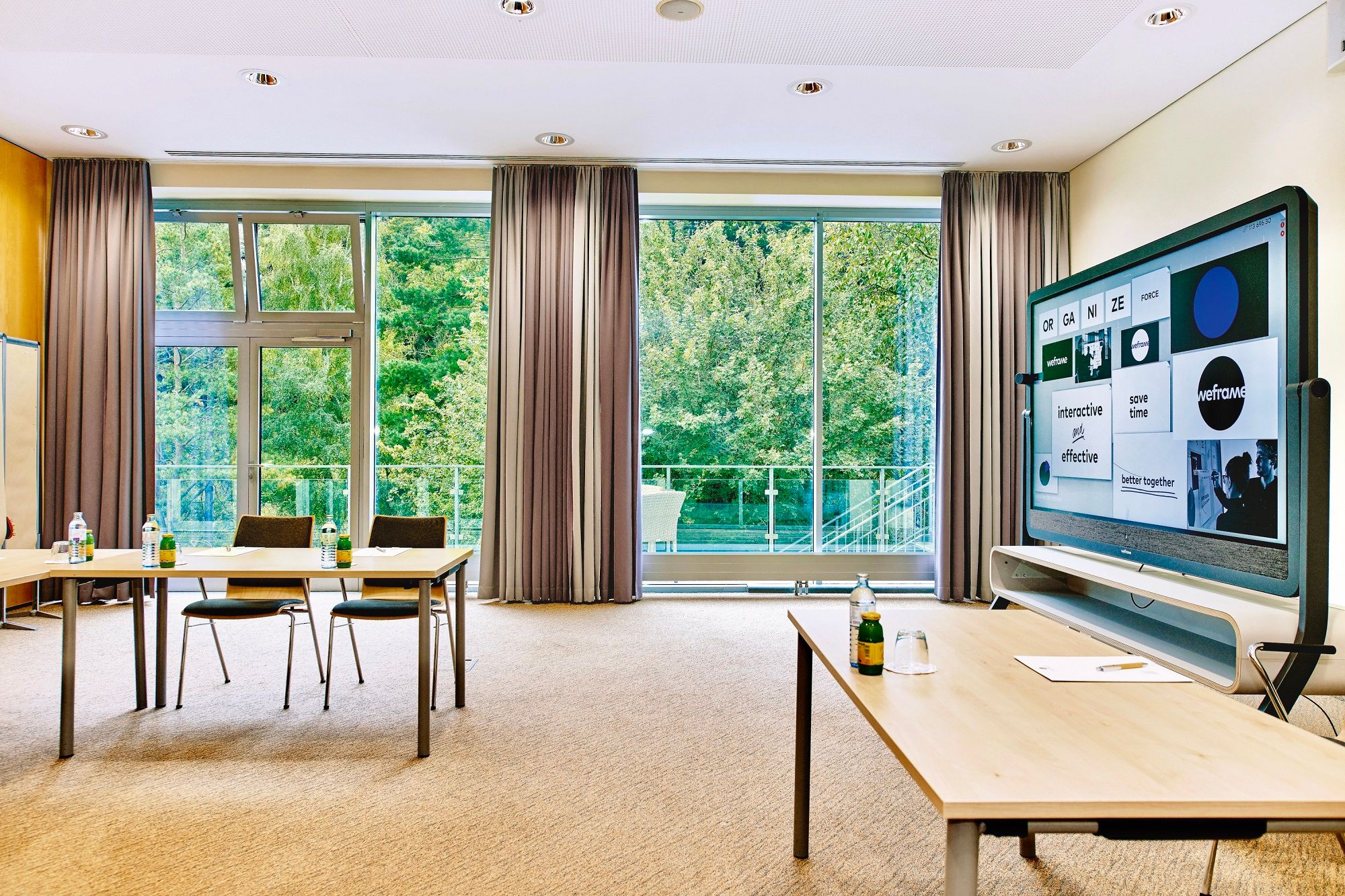 Moderner Seminarraum mit großen Fenstern, Tischen, Stühlen und einem interaktiven Bildschirm.