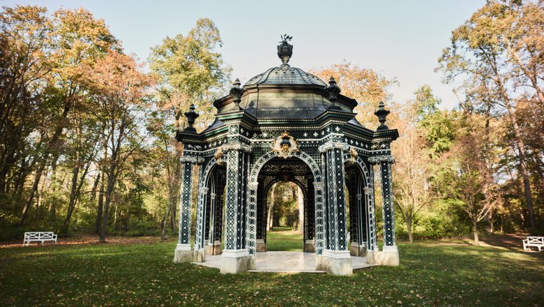 Ein kunstvoll gestalteter Pavillon im Gr&uuml;nen, umgeben von B&auml;umen im Herbst.