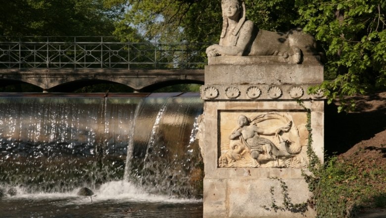 Steinfigur einer Sphinx neben einem Wasserfall und einer Br&uuml;cke im Park.