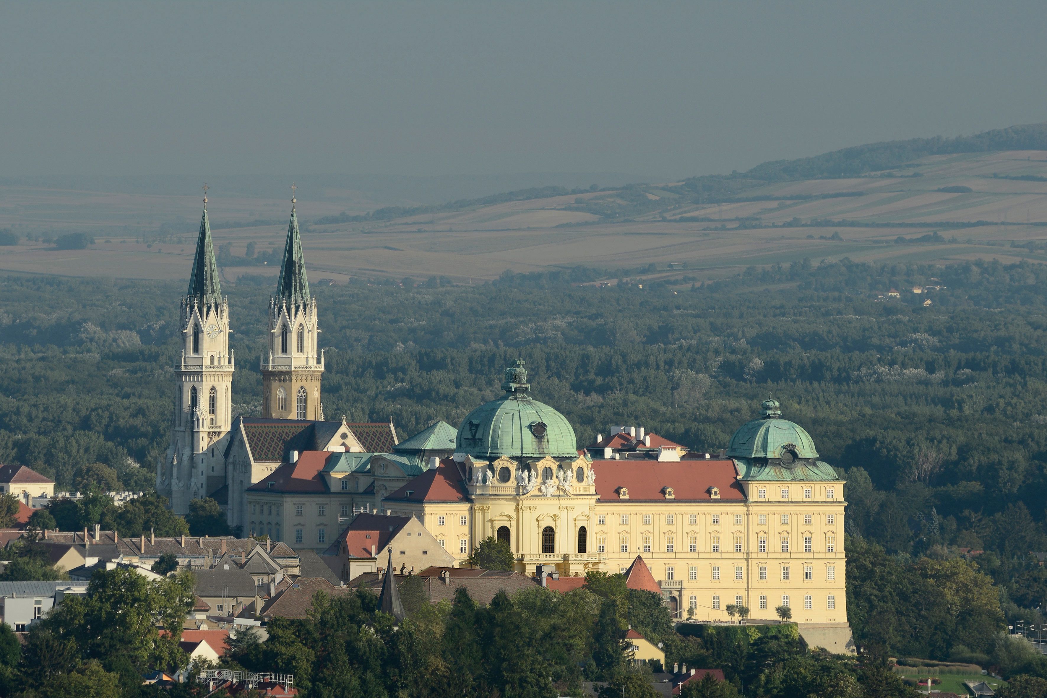 Luftaufnahme des Stifts Klosterneuburg mit zwei Türmen und einer Kuppel, umgeben von Wald und Feldern.