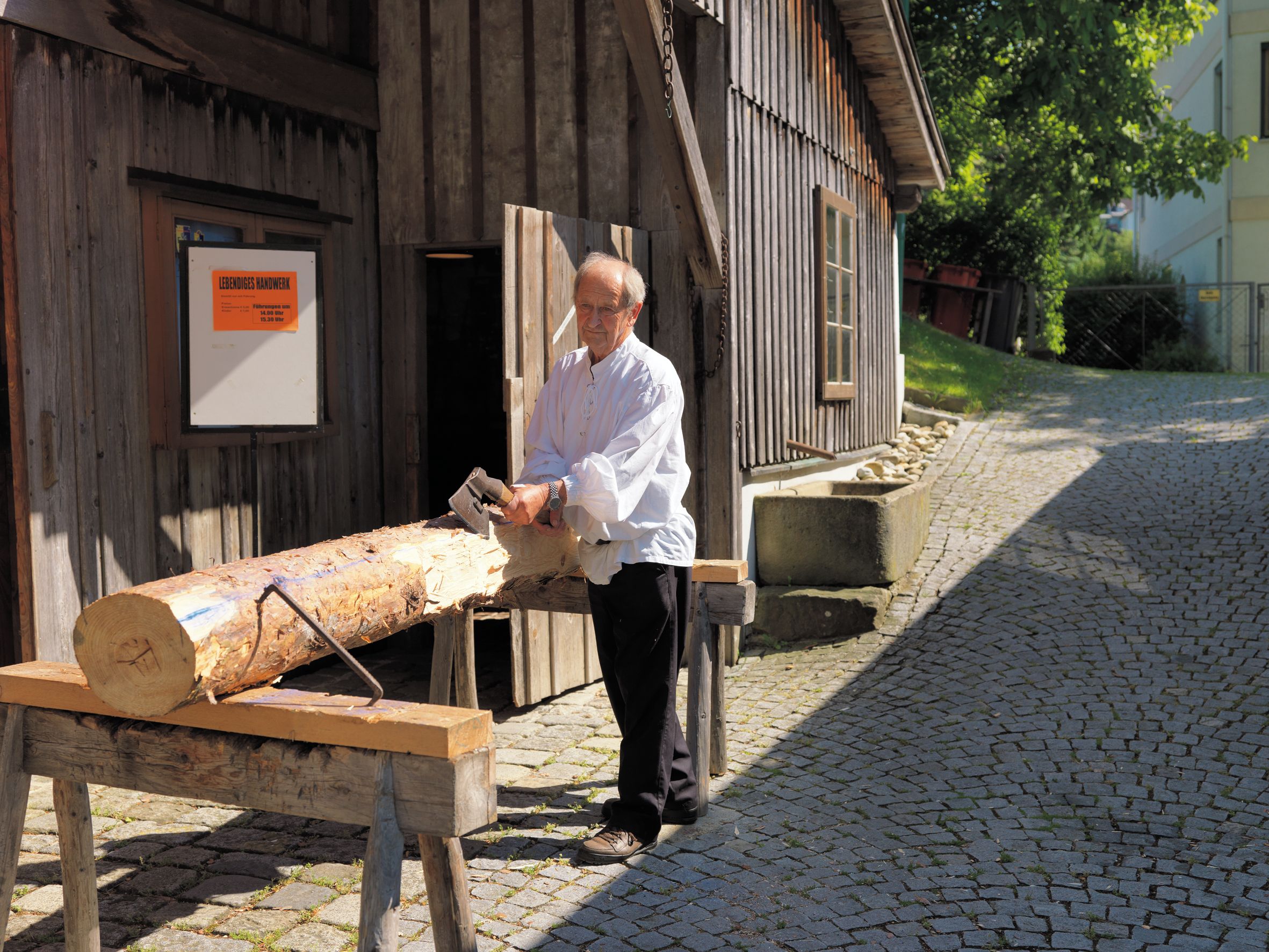 A man is working on a tree trunk with an axe in front of a wooden house.